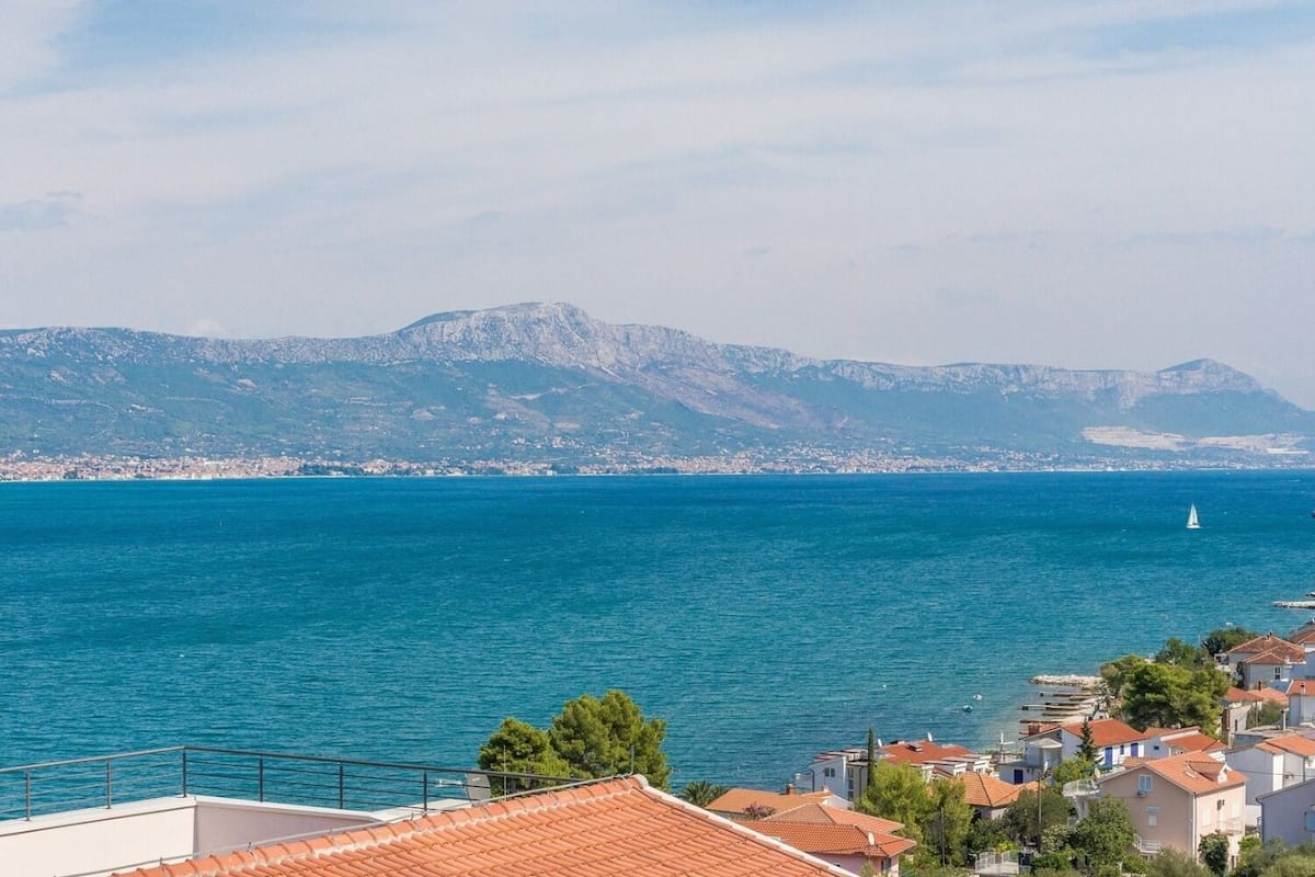 Une ville côtière aux bâtiments aux toits de tuiles rouges, bordée par une mer d’un bleu limpide et dominée par un paysage montagneux.