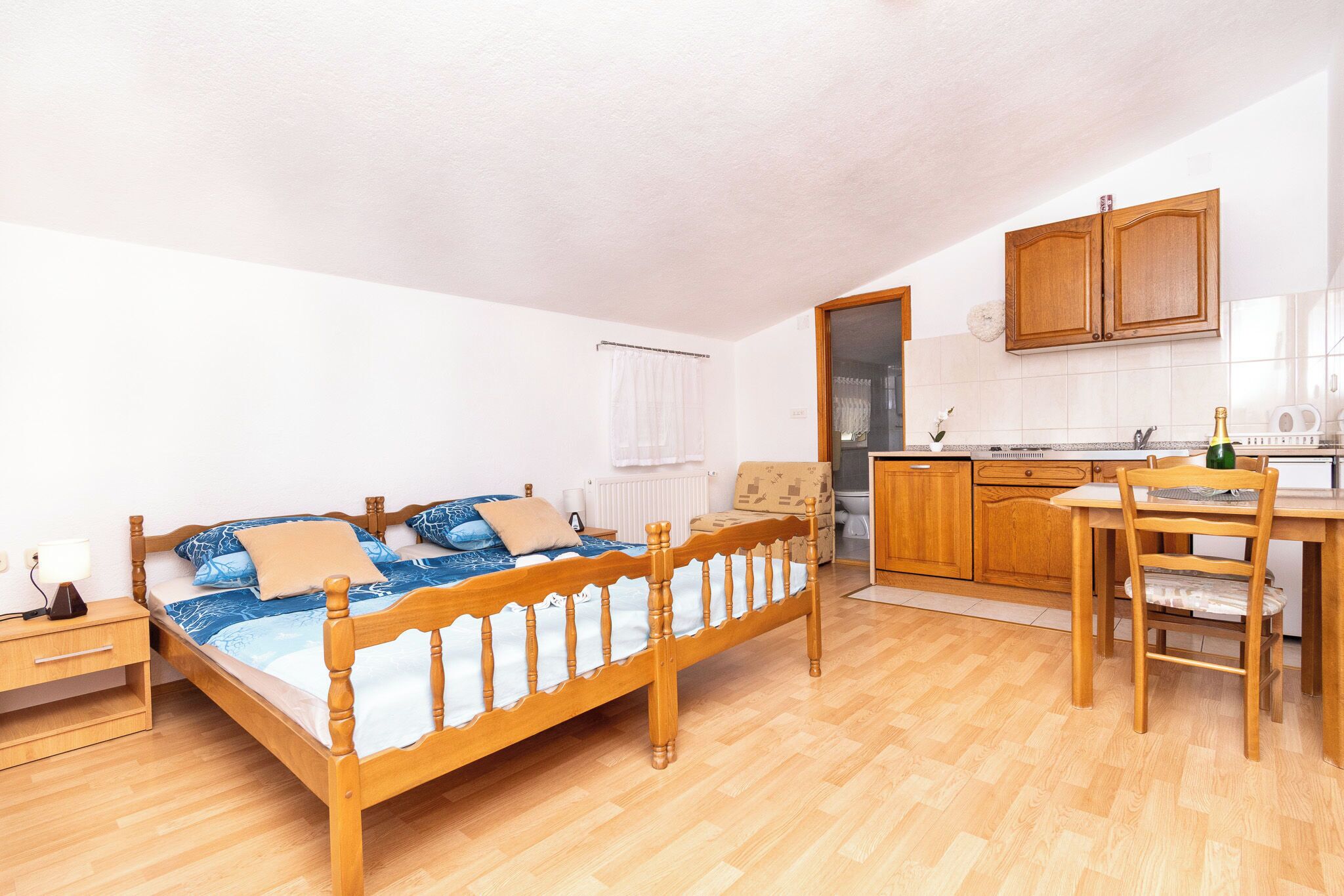 Een kamer met een houten bed, een klein bureau, een stoel, een kitchenette en een badkamer.