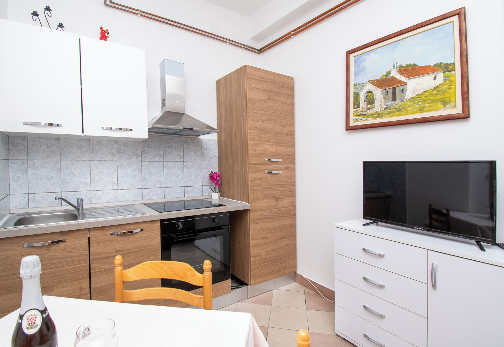A modern kitchen with white cabinets, a built-in oven, a wooden chair, and a TV on a white cabinet.