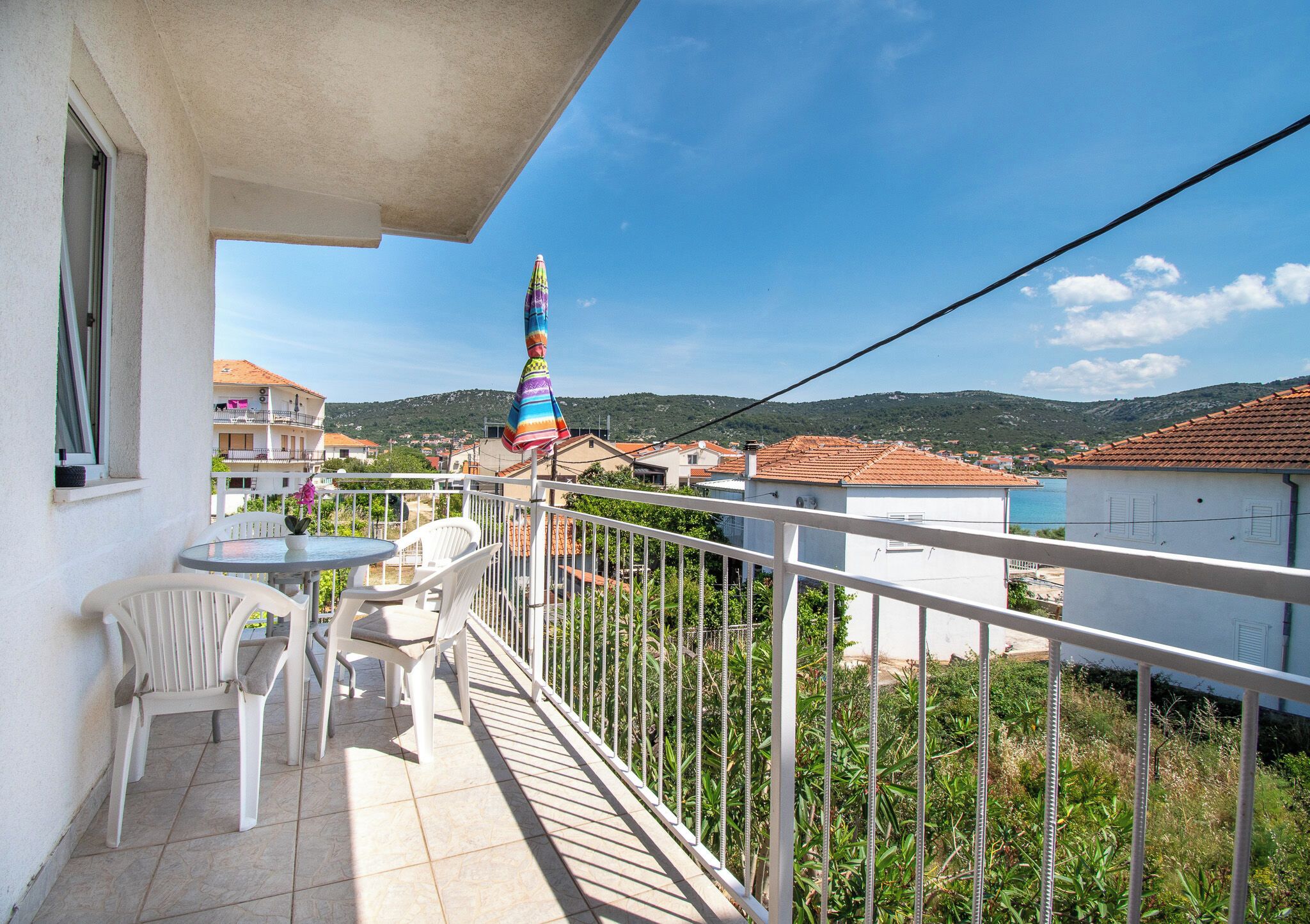 Ein Balkon mit Tisch und Stühlen, einem bunten Sonnenschirm und Blick auf eine Stadt und Hügel.