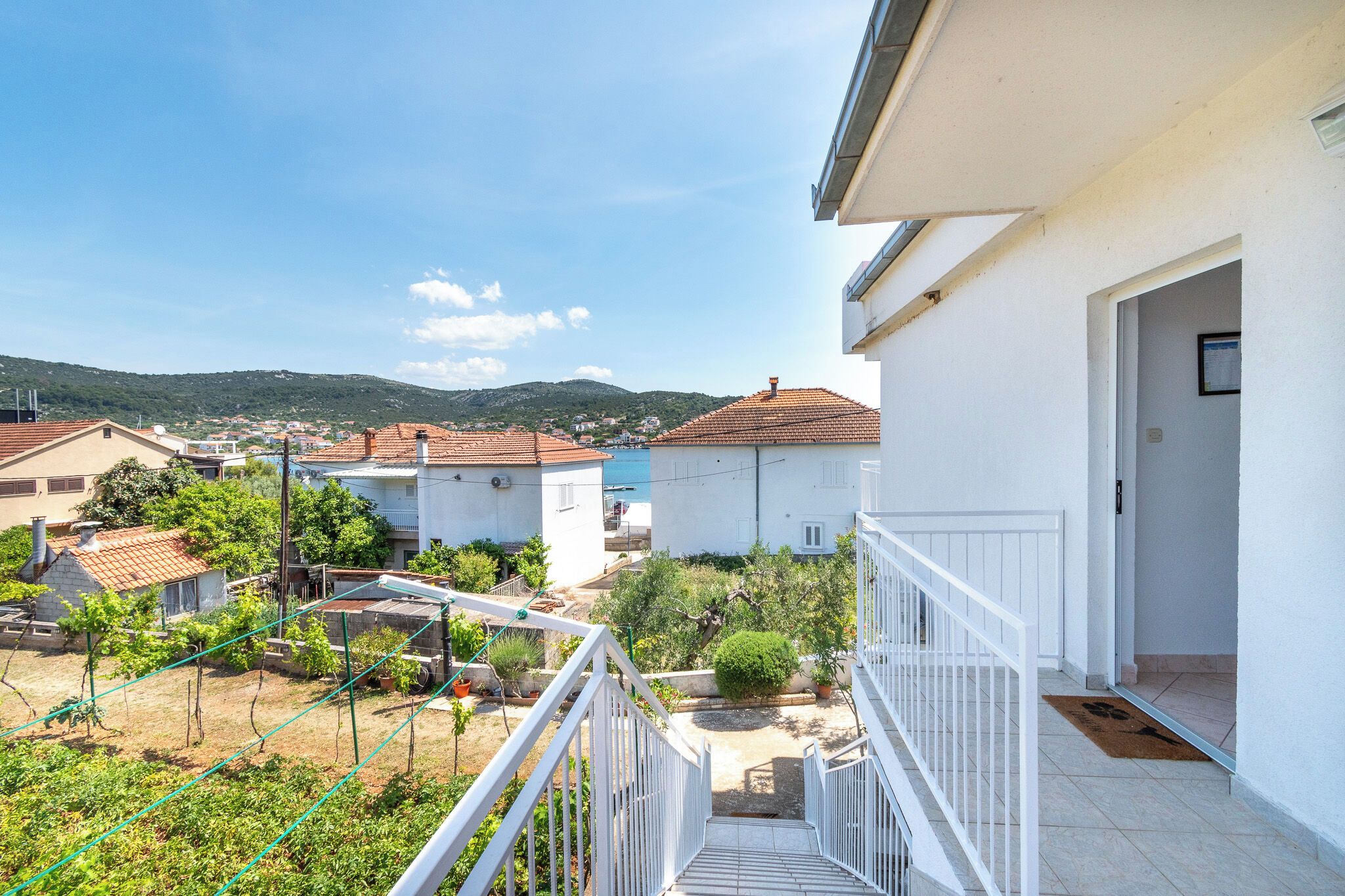 A residential area with a white building, a balcony with a white railing, and a view of houses and a body of water in the distance.
