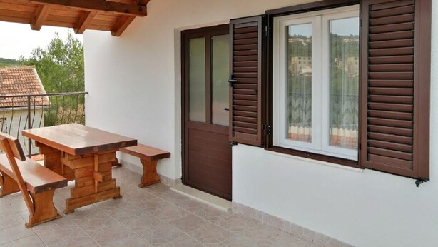 A wooden table and benches under a wooden ceiling, a door with brown shutters, and a window with a view of buildings. - Apartments Branko (Vis)