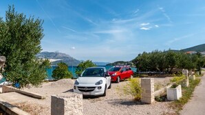 A parking area with a white car in the foreground and a red car in the background, with a scenic view of mountains and a body of water in the distance. - Apartments Zeljko (Korčula)