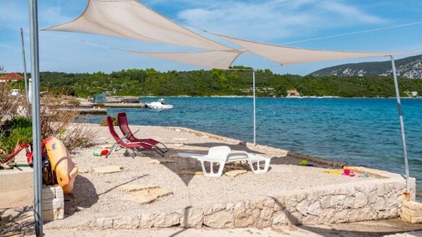 A beach with a stone platform, white plastic chairs, and a boat in the background. - Apartments Zeljko (Korčula)
