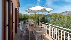 A balcony with a table and chairs, a white umbrella, and a view of a lake and mountains. - Apartments Zeljko (Korčula)