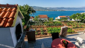 A terrace with a view of the sea, a garden, and a building with a tiled roof. - Apartment Marija (Trogir)