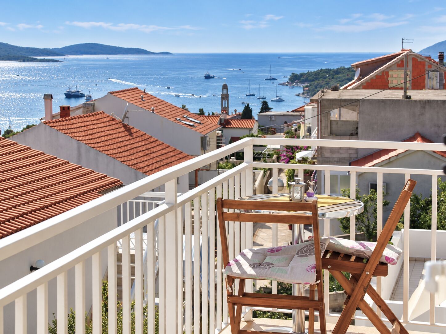 A balcony with a wooden chair and table, overlooking a coastal town with boats and buildings.