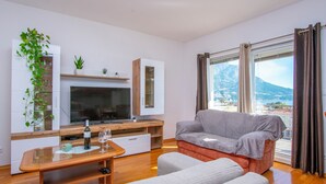 Egy modern nappali, melyben egy síkképernyős televízió, fából készült bútorok, egy üvegasztal található, és nagy ablakokon keresztül kilátás nyílik a hegyekre.