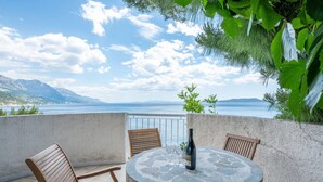 A round stone table with chairs, a bottle, and glasses on a balcony overlooking a body of water and mountains. - Apartments Marko (Omis)
