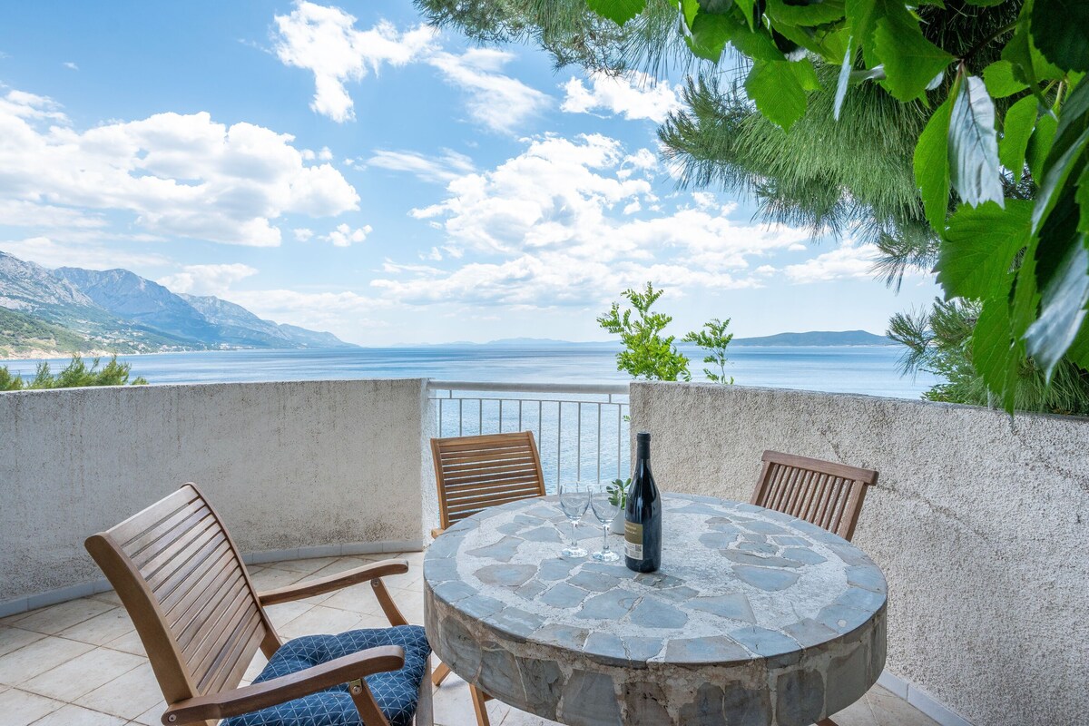 Een ronde stenen tafel met stoelen, een fles en glazen op een balkon met uitzicht op een waterpartij en bergen.