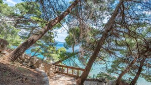A stone pathway leading to a wooden bench overlooking a body of water, surrounded by pine trees. - Apartments Marko (Omis)