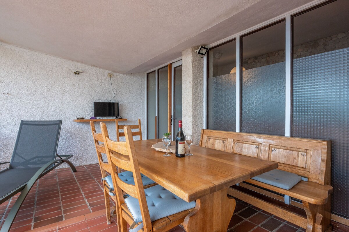 Een houten eettafel met stoelen en een bank, een televisie op een standaard, en een raam met gezandstraald glas.