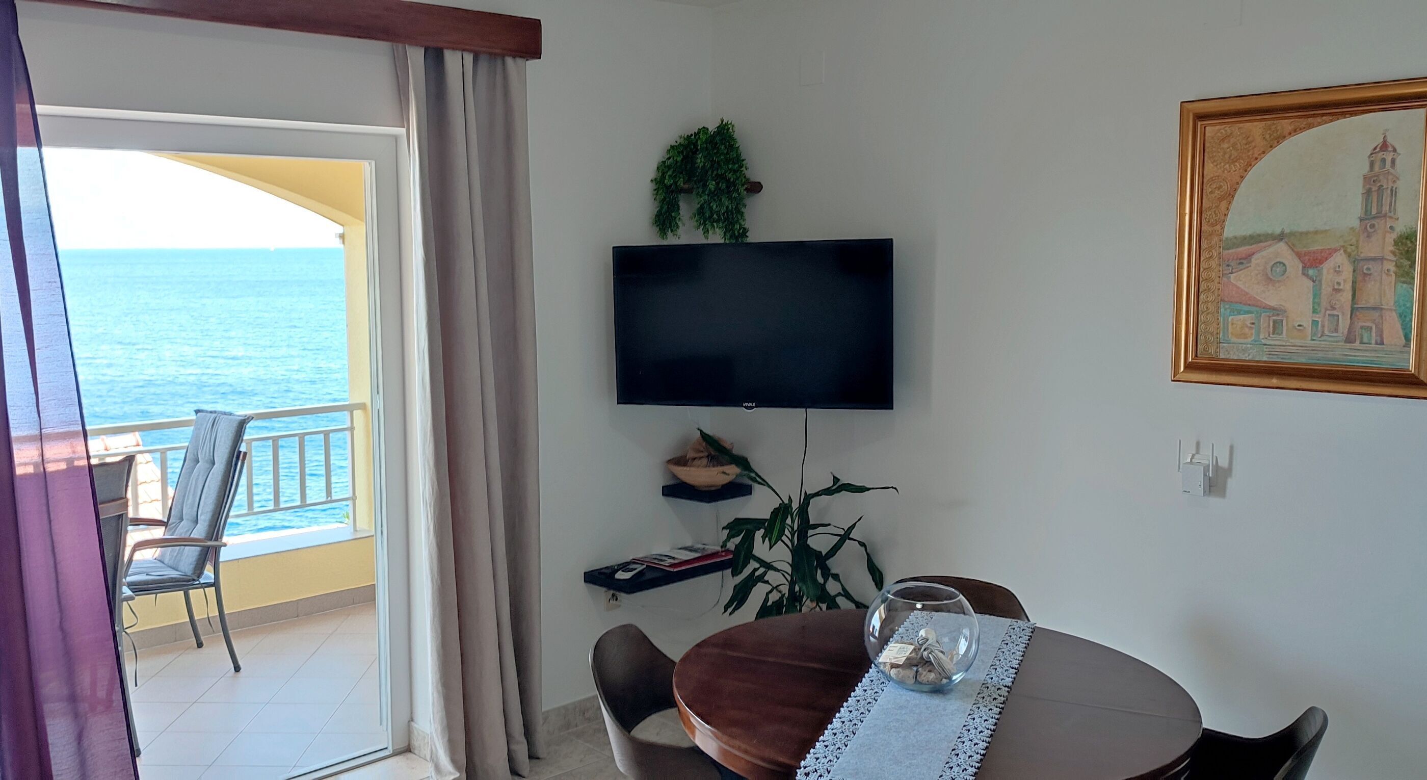 Una habitación con televisión, una mesa de comedor, sillas, una planta y vista al mar.