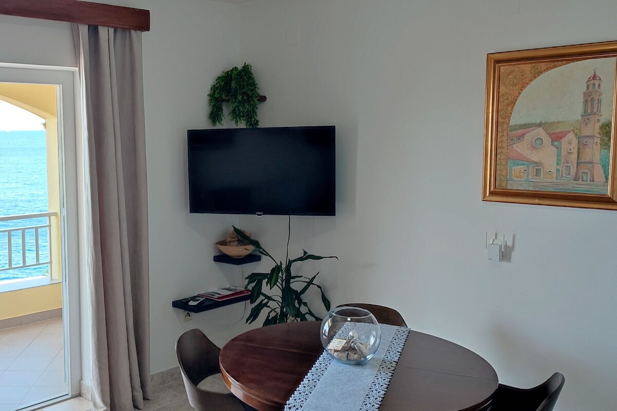 Huoneessa on seinälle asennettu televisio, ruokapöytä tuoleineen, huonekasvi ja taulu.