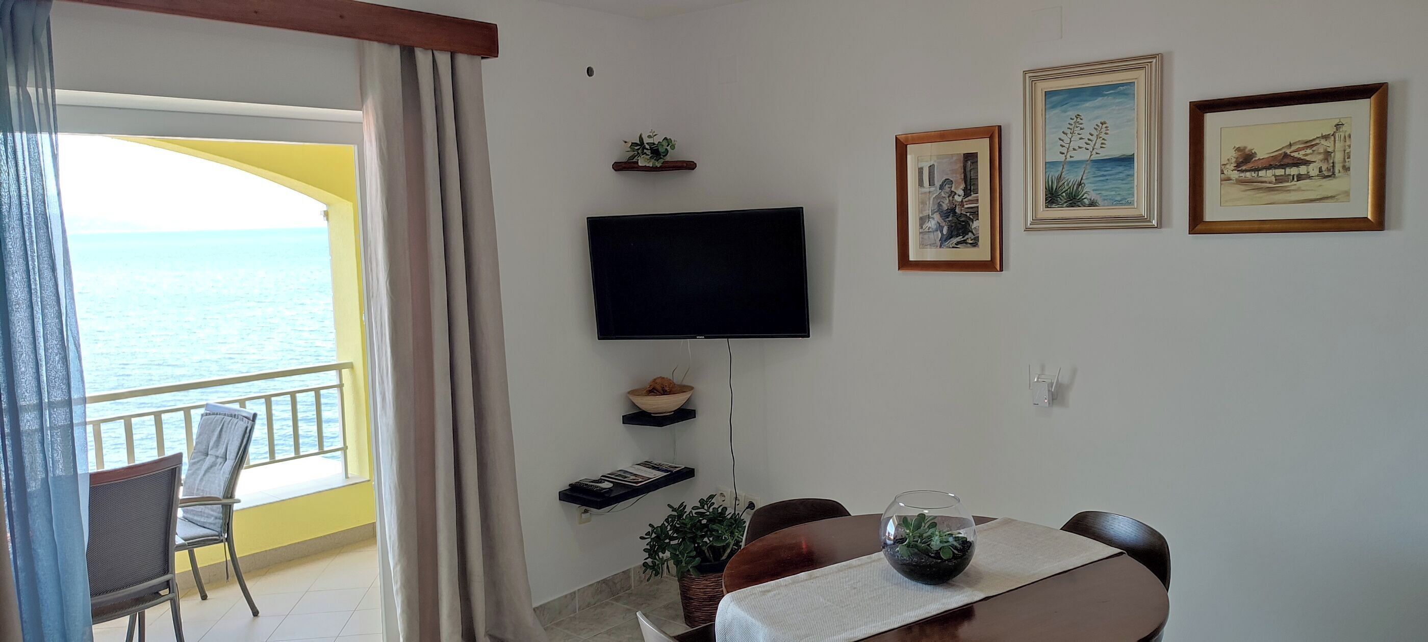 Habitación con televisión, obras de arte enmarcadas, mesa de comedor con sillas, planta en maceta y vista al mar.