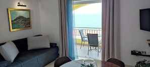 Una habitación con sofá, una mesa con un vaso, una silla, televisor y balcón con vista al mar.