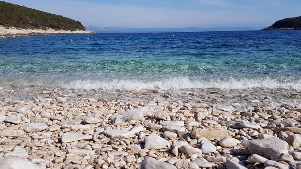 Una spiaggia ciottolosa con acque limpide e colline sullo sfondo.