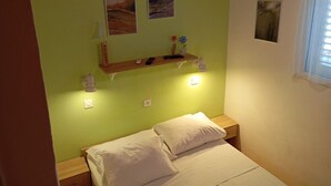 A bedroom with a bed, two wall-mounted shelves with decorative items, and a window with blinds. - Apartments Ivo (Korčula)