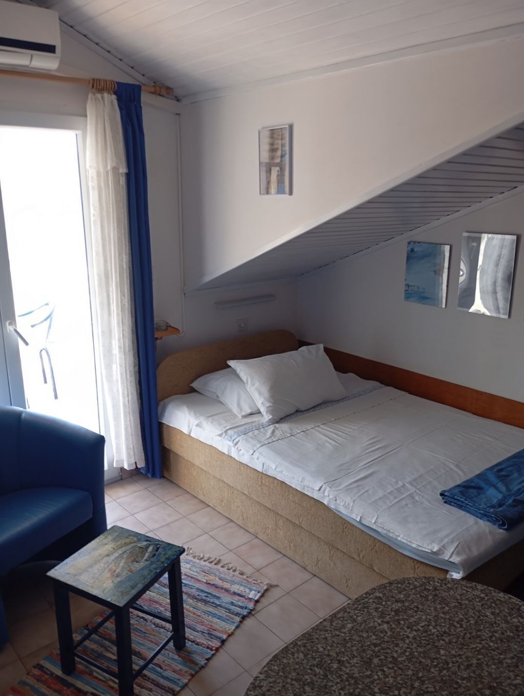 A single bed with white bedding, a blue chair, a small table, and a patterned rug in a room with a sloped ceiling and a window with curtains.
