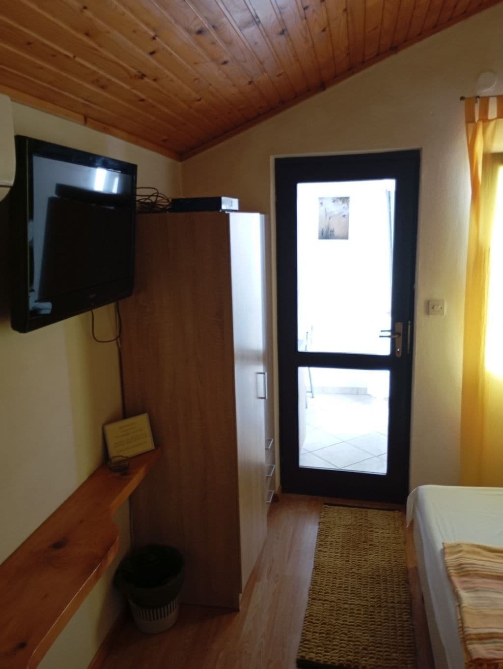 A room with a wooden ceiling, a television mounted on the wall, a wardrobe, a door leading outside, and a bed with a striped blanket.