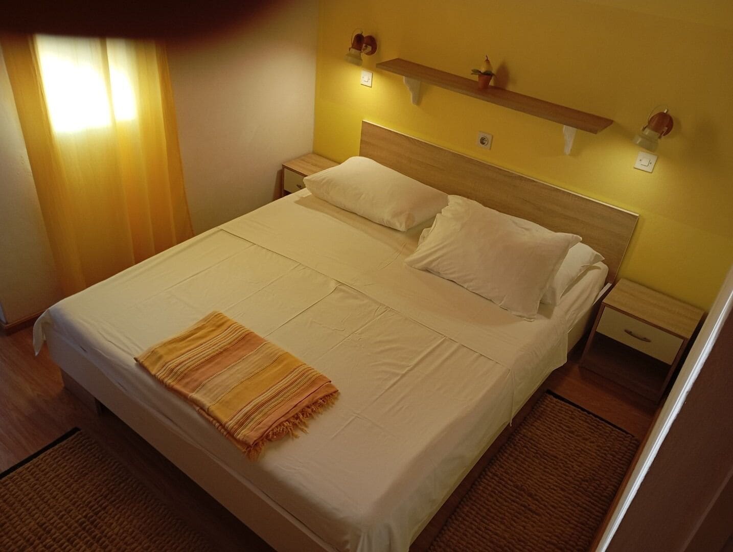 Een net opgemaakt bed met witte lakens en een gestreept plaids, een houten hoofdbord, nachtkastjes met lampen, en een plank met decoratieve objecten.