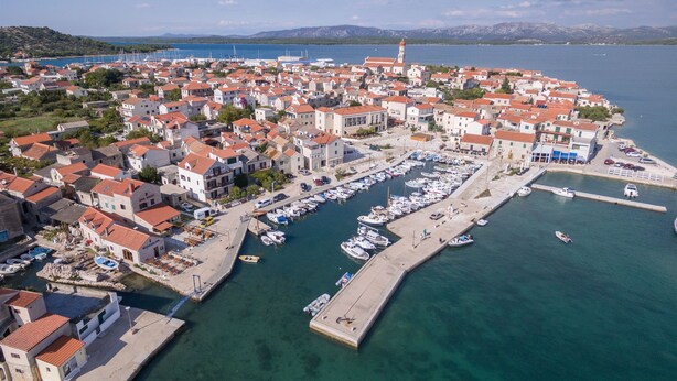 An aerial view of a coastal town with a marina, houses with red-tiled roofs, and a prominent church with a bell tower. - Apartment Mili Sea View Betina (Tisno)