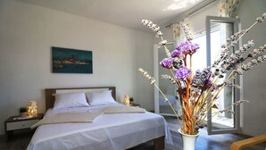 A neatly made bed with white linens, a bedside table with a lamp, and a vase with dried flowers on a wooden floor. - Apartment Mili Sea View Betina (Tisno)