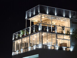 A multi-story building with large glass windows and balconies, featuring outdoor plants and interior lighting. - THE SARAI (Udaipur)