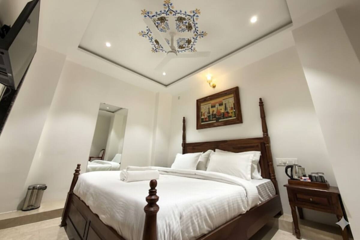 A hotel room with a wooden bed, a ceiling fan, a painting on the wall, and a small table with a kettle.