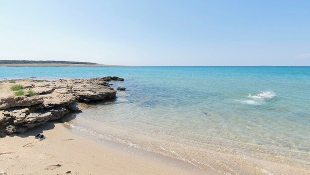 Egy tiszta vizű strand, sziklás képződményekkel, és egy pár cipő a homokban.