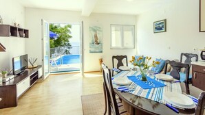A dining area with a table set for four, a TV on a stand, and a view of a balcony with a pool. - House Ned (Tucepi)