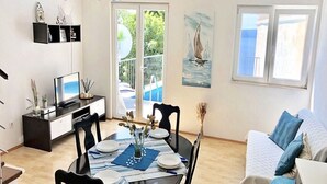 A dining area with a round table, chairs, and a TV on a stand. There is a balcony with a pool and a sailboat painting on the wall. - House Ned (Tucepi)
