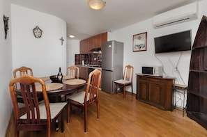 A dining area with a wooden table and chairs, a kitchen with wooden cabinets and appliances, and a mounted television. - Apartments Ema (Murter-Kornati)