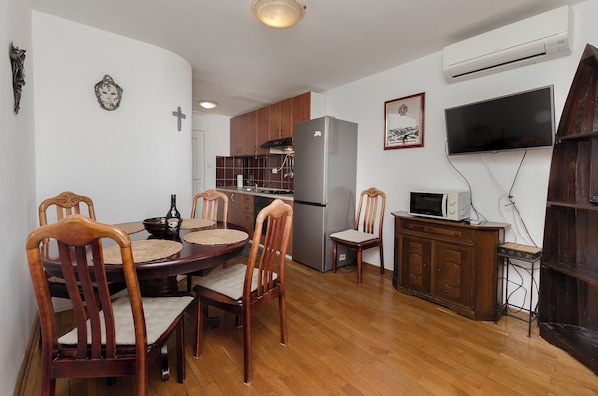 A dining area with a wooden table and chairs, a kitchen with wooden cabinets and appliances, and a mounted television. - Apartments Ema (Murter-Kornati)