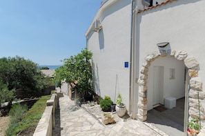 A white building with a stone archway, a small garden with potted plants, and a view of the sea. - Apartments Ema (Murter-Kornati)