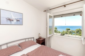 A bedroom with a bed, a nightstand, a lamp, a window with a view of the sea, and a painting on the wall. - Apartments Ema (Murter-Kornati)