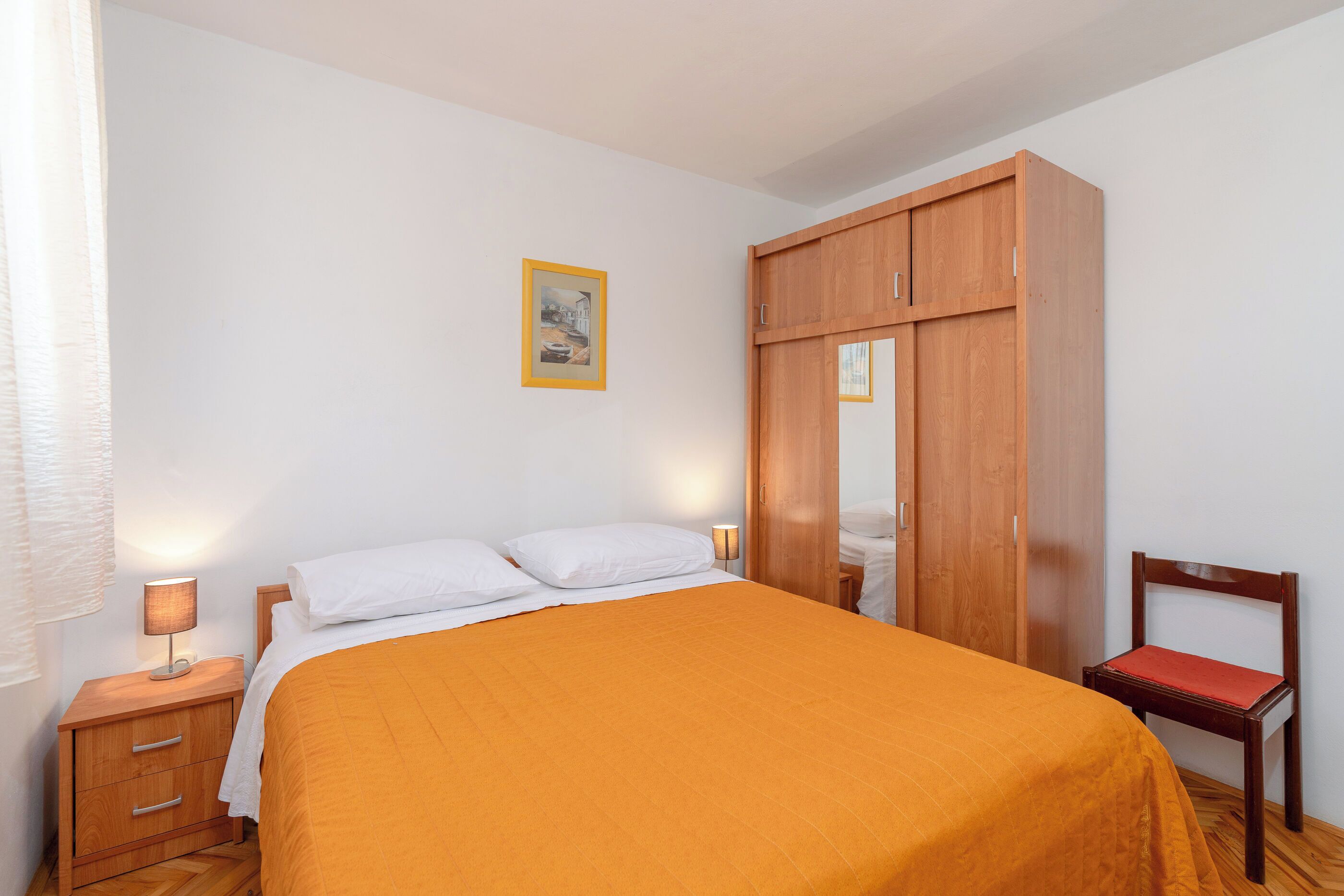 A bedroom with a bed, wooden wardrobe, bedside table, and a chair.