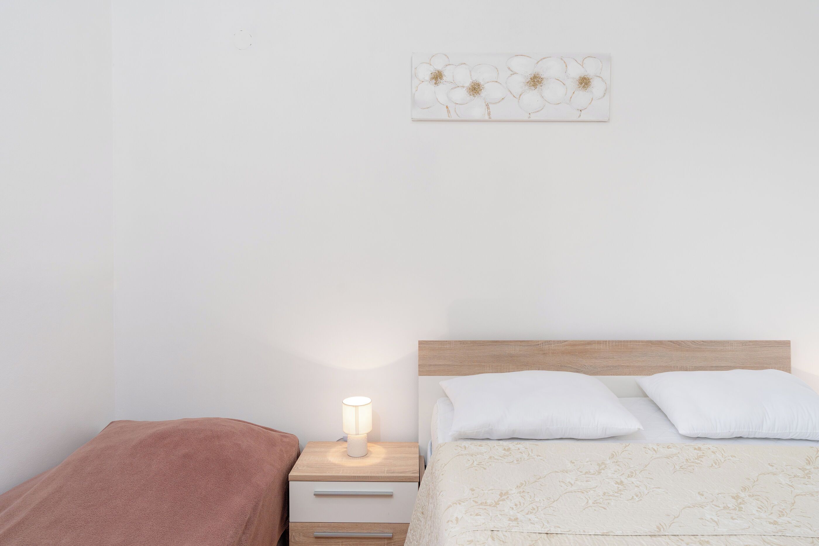 A bedroom with a wooden headboard, a bedside table with a lamp, a brown towel, and a floral artwork on the wall.