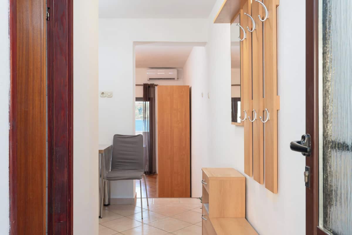 Un pasillo con una puerta de madera, una silla gris, un armario de madera y un espejo con marco metálico.