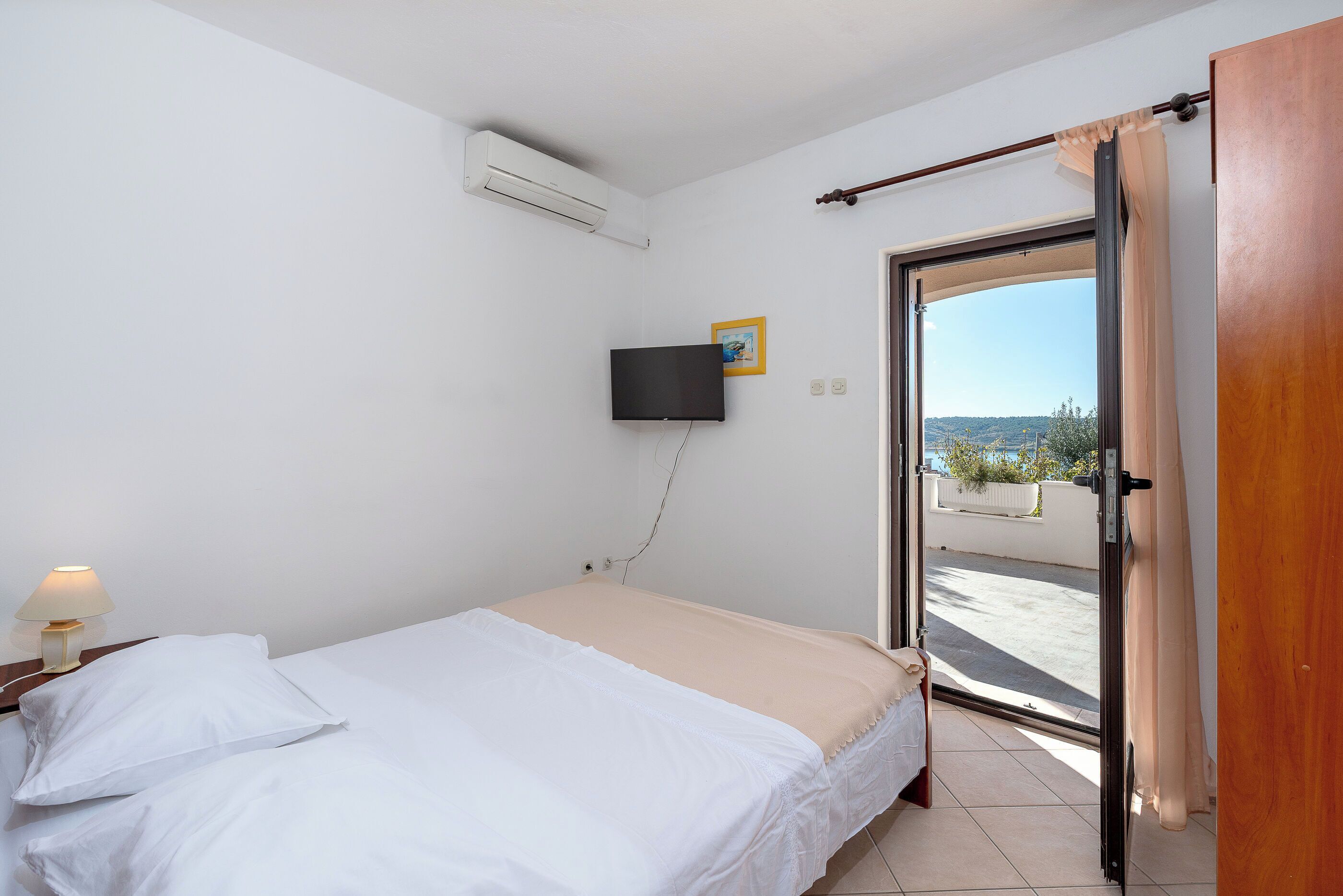 A bedroom with a bed, a TV mounted on the wall, a window with curtains, and an air conditioning unit.