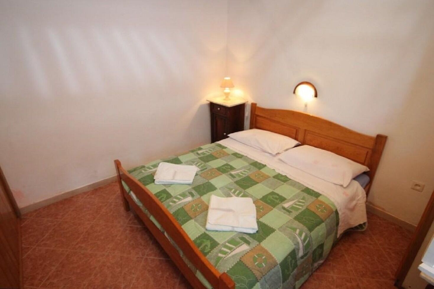 A single bed with a wooden headboard, a green and white patterned bedspread, and two white pillows. There is a nightstand with a lamp on it to the left of the bed.