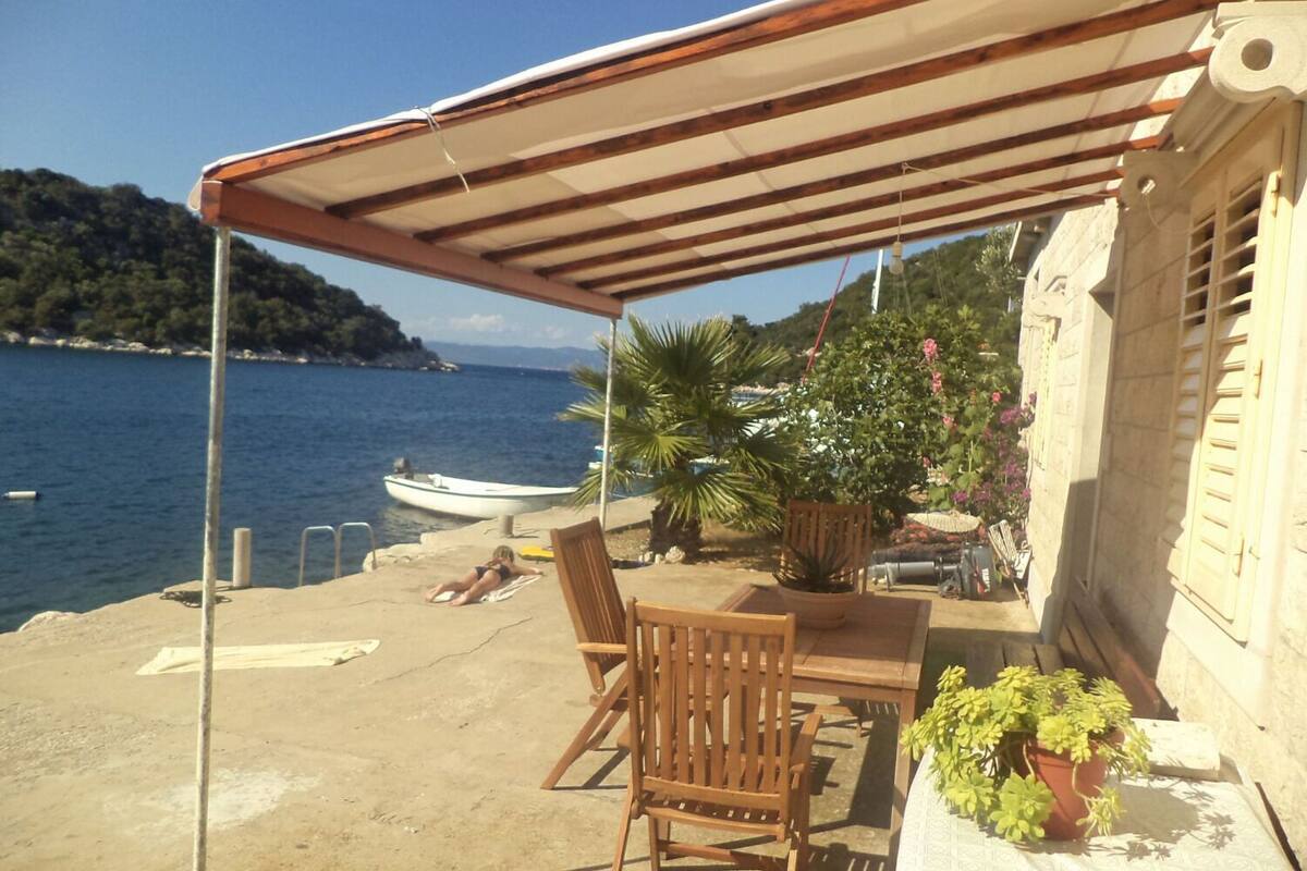 A covered outdoor seating area with a table and chairs, overlooking a body of water with a boat and a mountainous landscape in the background.