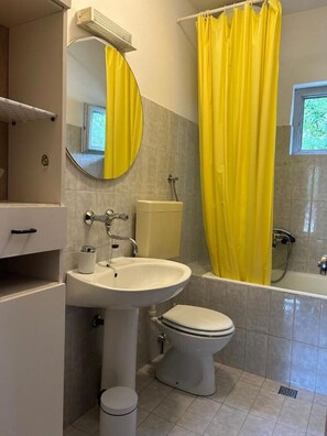 Ein Badezimmer mit Toilette, Waschbecken und Badewanne, mit einem gelben Duschvorhang und einem Fenster.