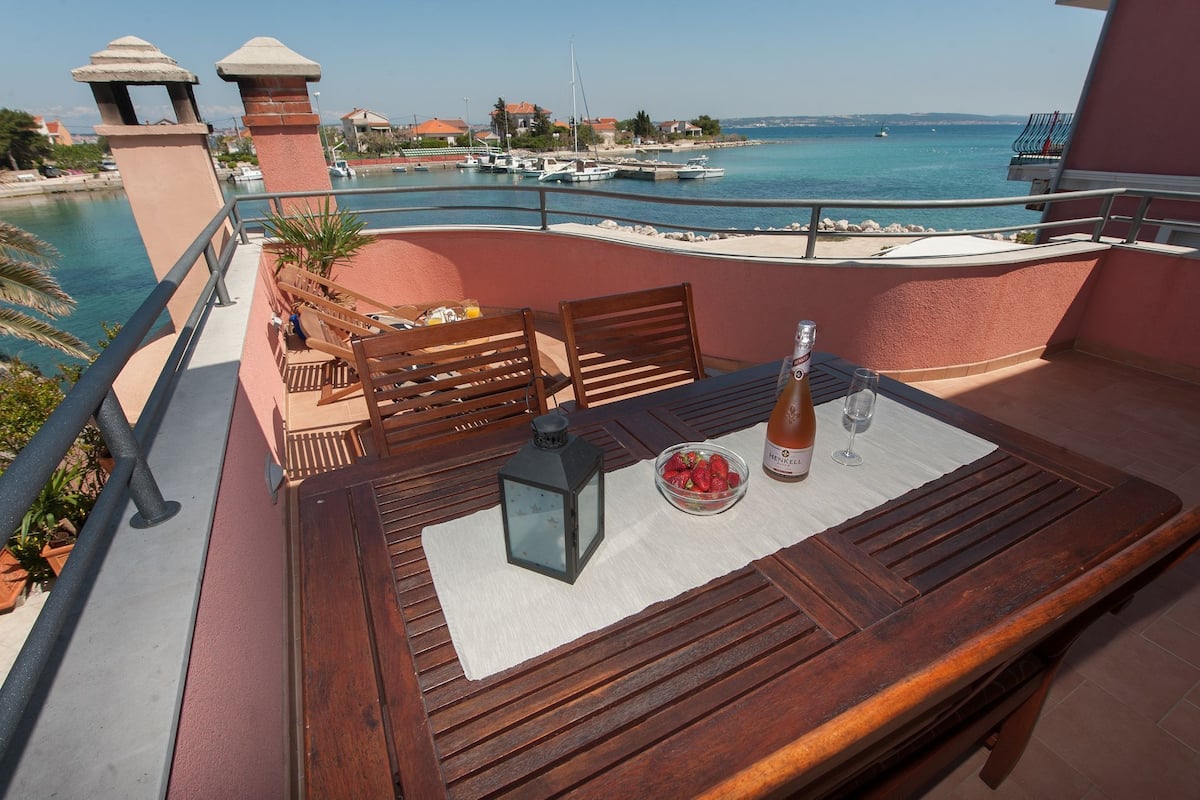 A balcony with a wooden table, chairs, a bottle of Henkell beer, and a glass. The balcony overlooks a marina with boats and houses in the background.