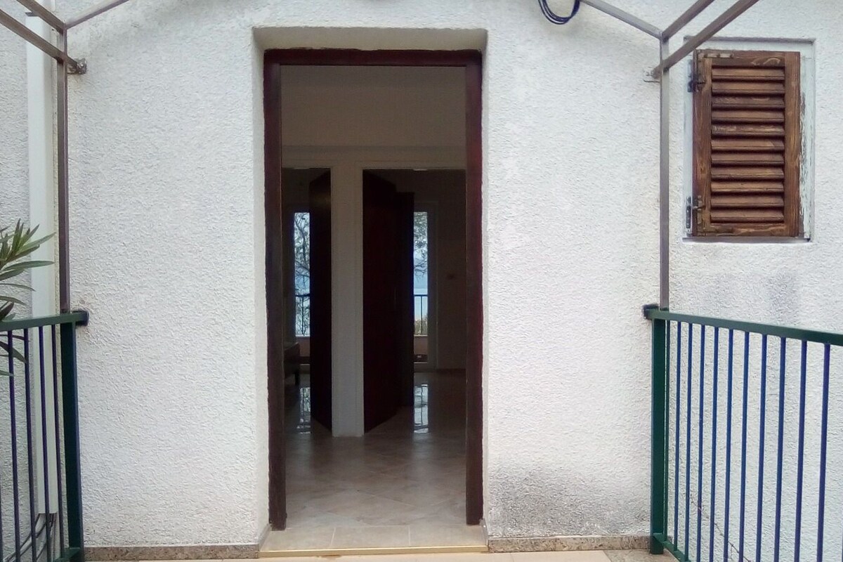 Une entrée d’immeuble avec une porte en bois, une petite fenêtre avec des volets et une rambarde en métal.