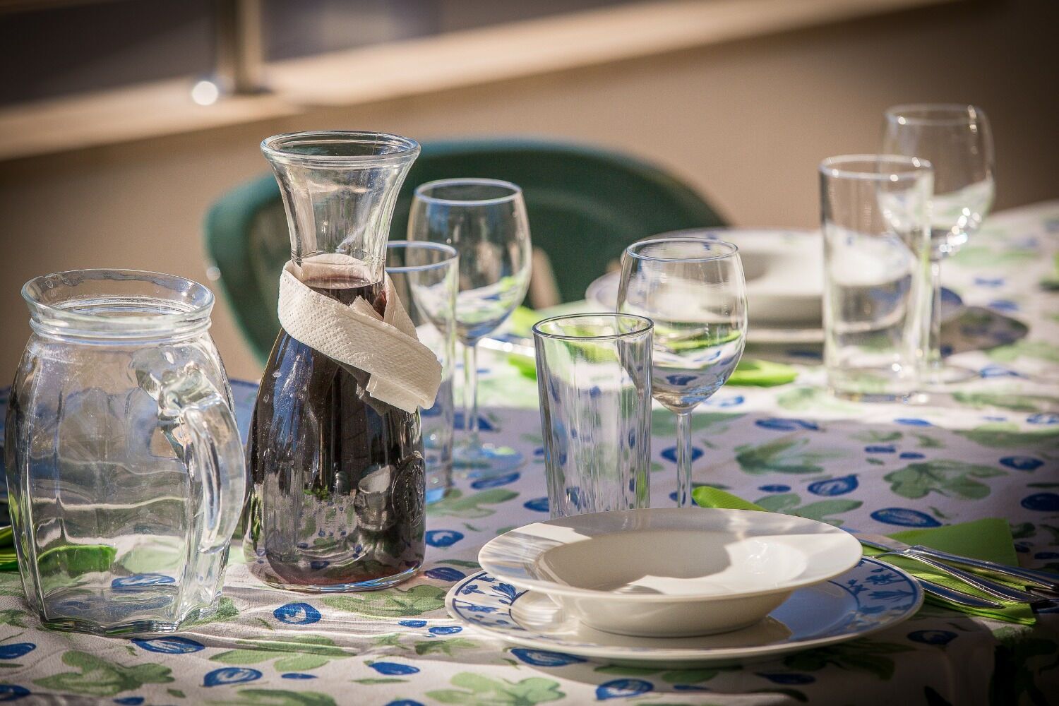 A table set with clear glassware, a bottle with a cloth, and a floral tablecloth.