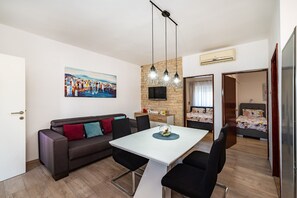 A modern living room with a sofa, dining table, and chairs. There is a painting on the wall, a TV, and a bed in the adjacent room. - Apartments Dreamy (Nin)