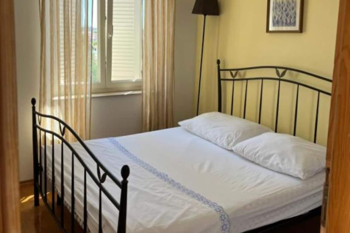 Un lit simple avec un cadre en métal, une fenêtre avec des rideaux, une lampe sur pied et un tableau encadré au mur.