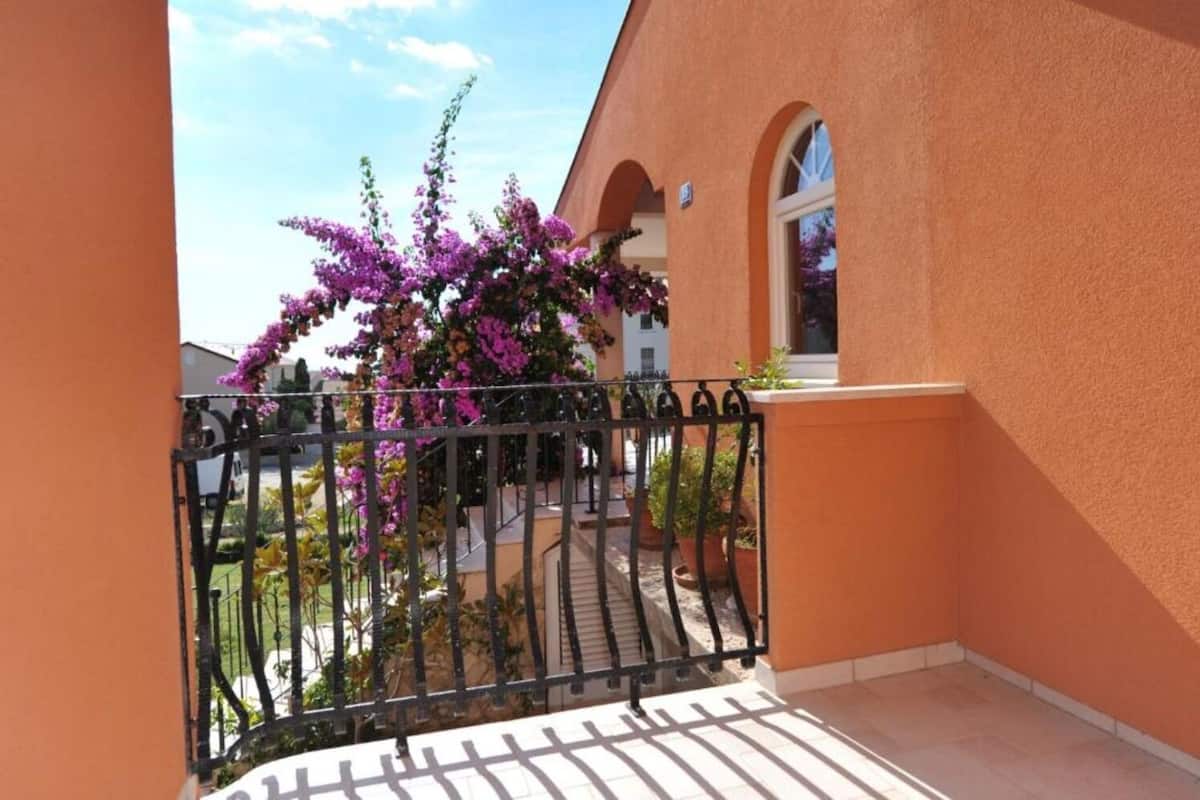 Un balcon orné d’une balustrade en fer forgé, d’un pot de fleurs et d’un arbuste à fleurs violettes.
