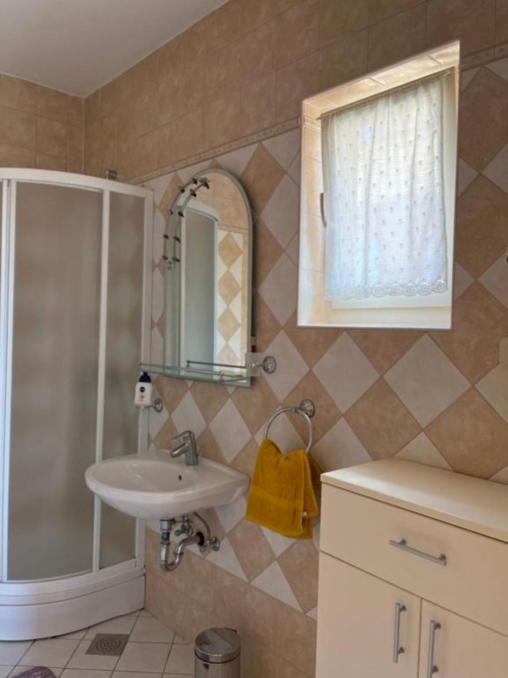 A bathroom with a shower, sink, mirror, and a window with curtains.